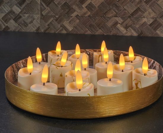 Een rond gouden dienblad bevat verschillende LE led theelichtjes 3D brandtijd 175 uur oplaadbaar - met Afstandsbediening - 6 Stuks - timerfunctie, gerangschikt op een donker aanrecht met een tegelachterwand met textuur op de achtergrond.