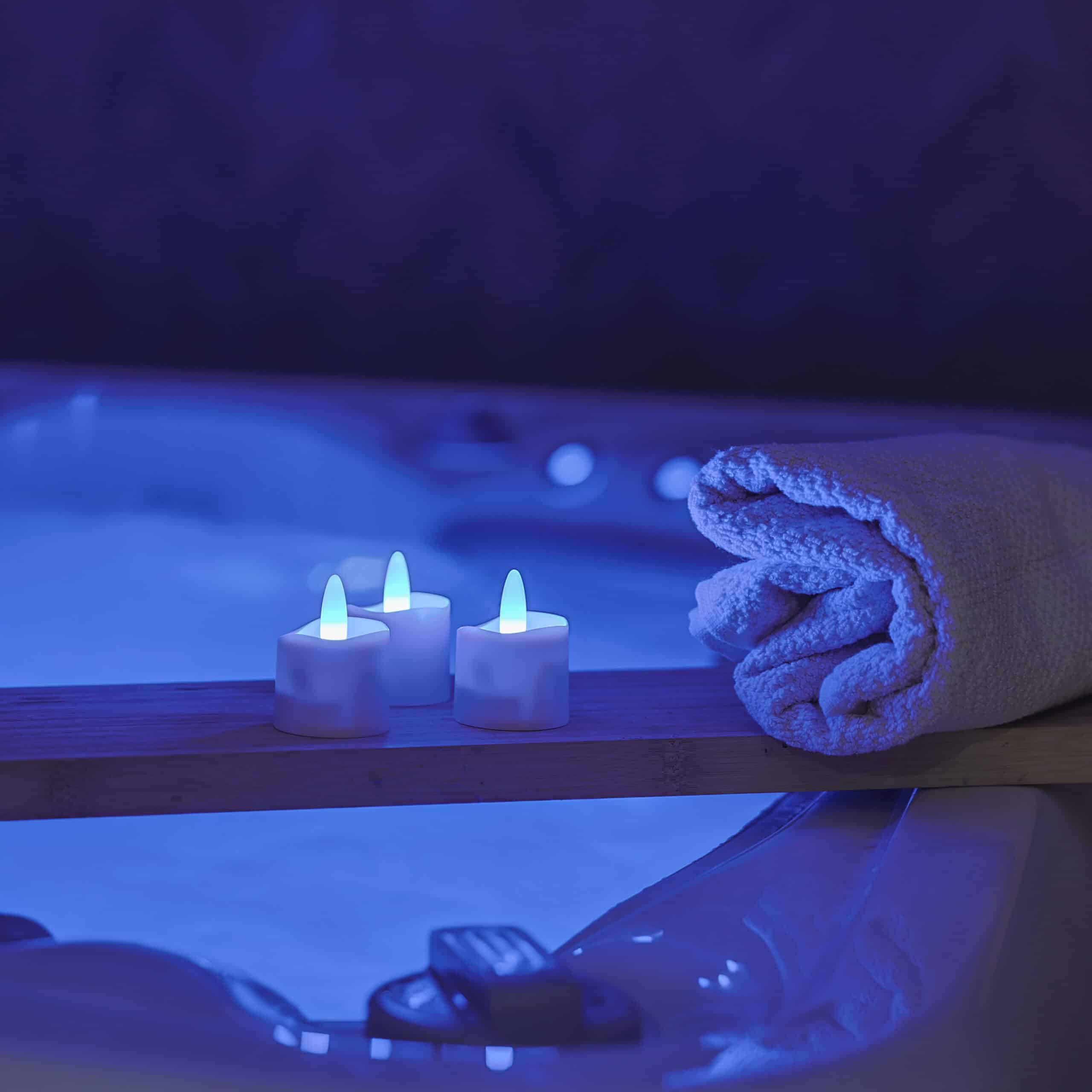 Een opgerolde handdoek en drie RGB LED-Waxine/theelichtjes oplaadbaar waterdicht (12 stuks) met afstandsbediening zitten op een houten plank boven een blauw verlicht bad en creëren een rustige, spa-achtige sfeer.