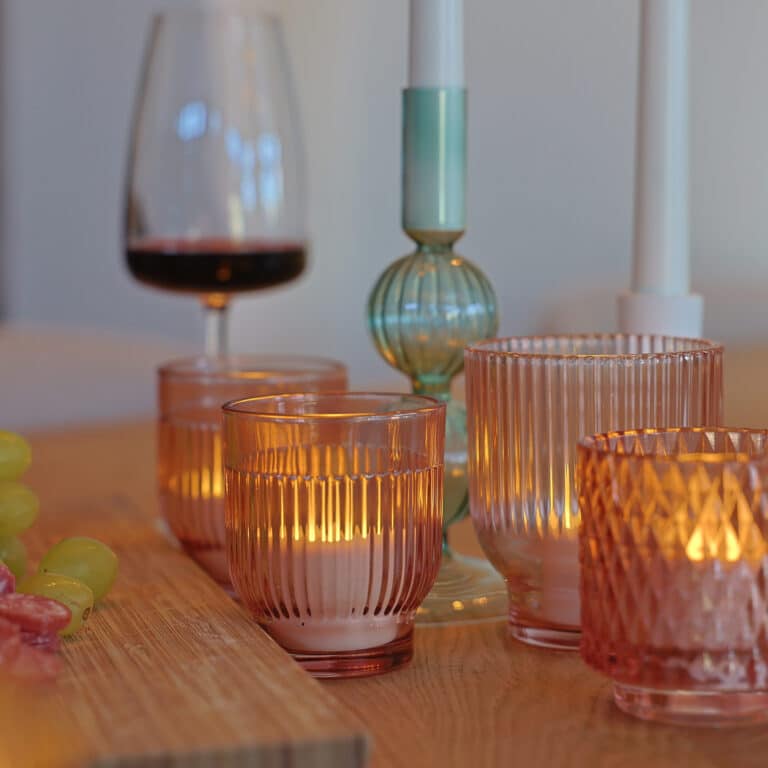 Een houten gedekte tafel met 12 waterdichte, oplaadbare LED waxinelichtjes (175 uur brandtijd, warm wit) met afstandsbediening, een glas rode wijn, witte en turquoise kandelaars en een bord groene druiven en gesneden salami in de hoek.