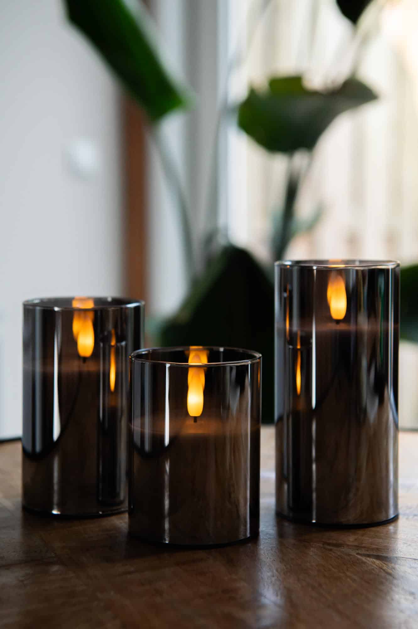 Three DORSETTO oplaadbare kaars in glas met 3D vlam staan op een houten oppervlak, hun sfeervolle licht weerkaatst in het glas. Op de achtergrond zorgen planten en daglicht voor de perfecte sfeer. Inclusief afstandsbediening, tot 105 uur brandtijd.