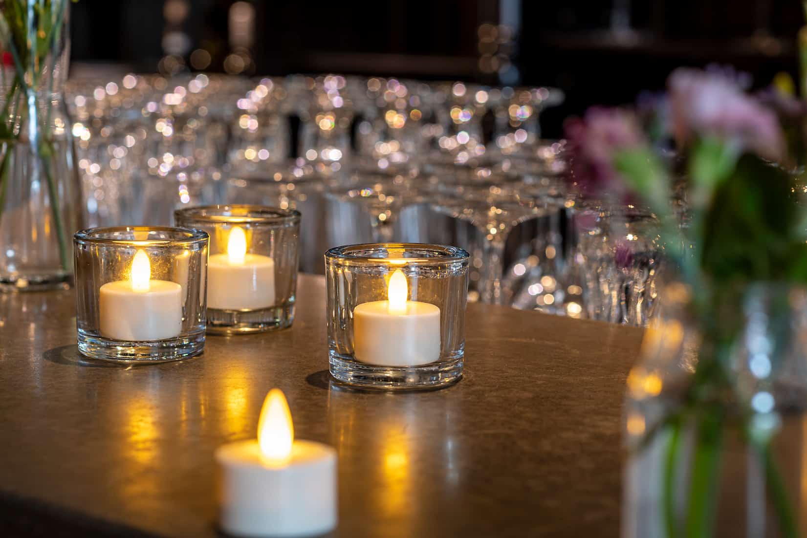 Twaalf Led waxinelichtjes waterdicht oplaadbaar brandtijd 175 uur warm wit met afstandsbediening staan in heldere houders op een aanrecht, met onscherp glaswerk en bloemen op de achtergrond, waardoor een warme, uitnodigende sfeer wordt gecreëerd.